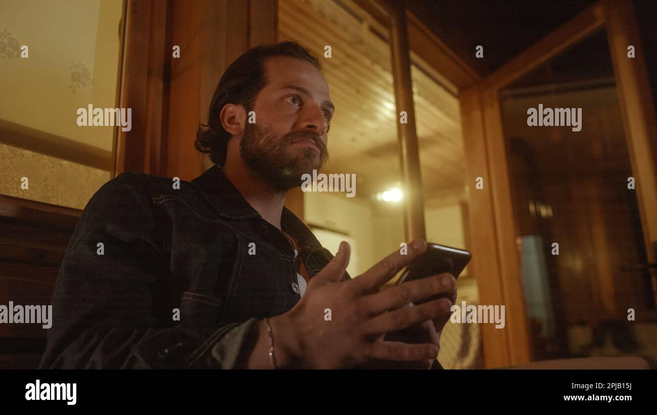 Er schrieb auf seinem Handy vom Balkon eines hölzernen Hauses, das von warmem gelben Licht beleuchtet wurde Stockfoto