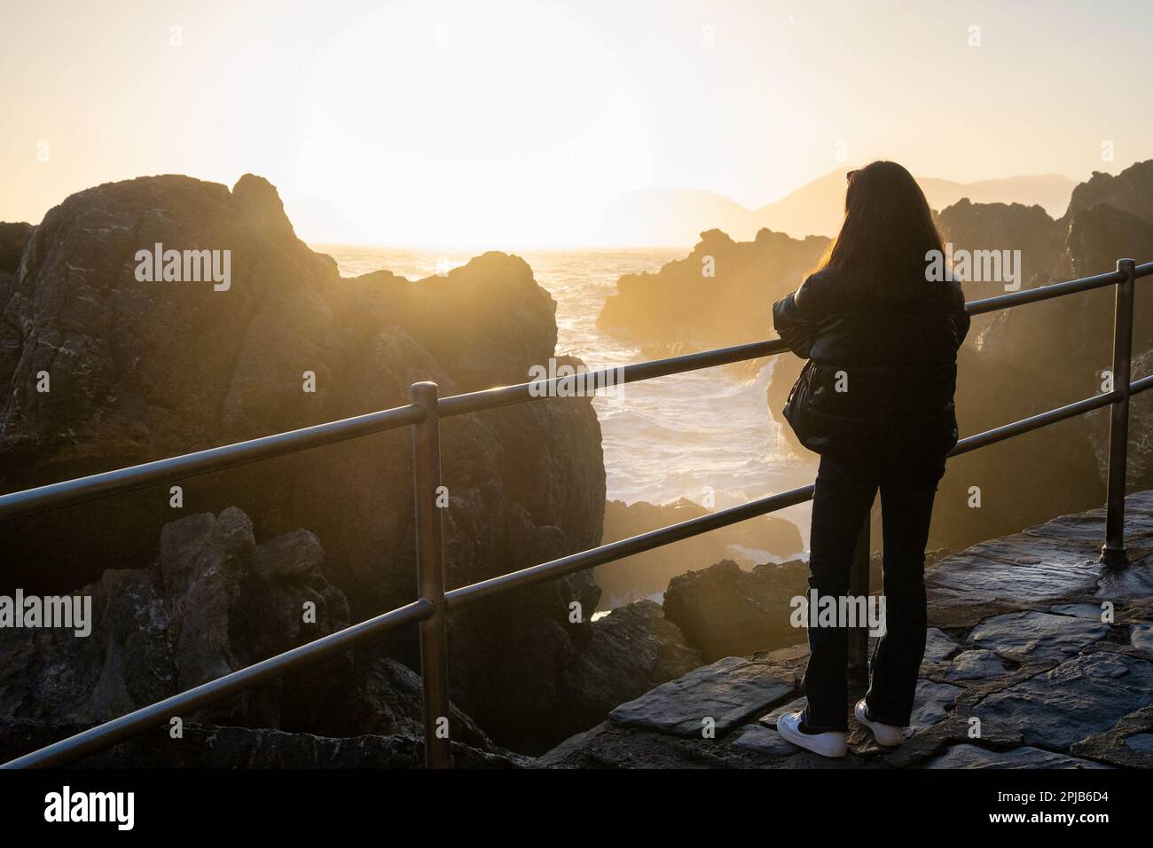 Eine einsame junge Frau steht nahe am Meer und beobachtet die Wellen bei Sonnenuntergang. Silhouette. Tourist. Raue See. Blick von hinten Stockfoto