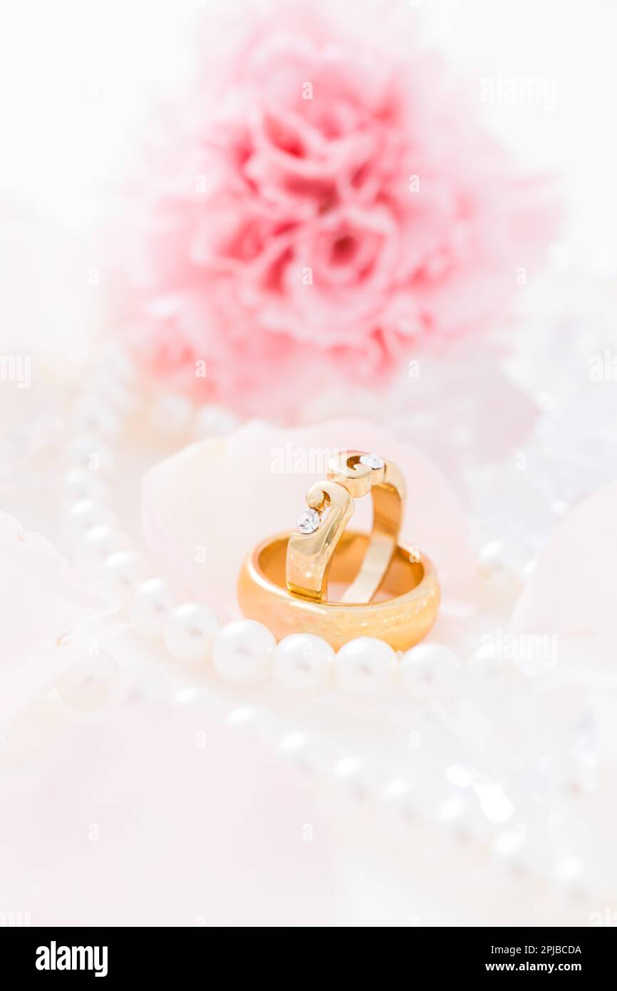 Goldene Eheringe mit Perlenkette mit rosa Blume im Hintergrund, weicher Fokus Stockfoto
