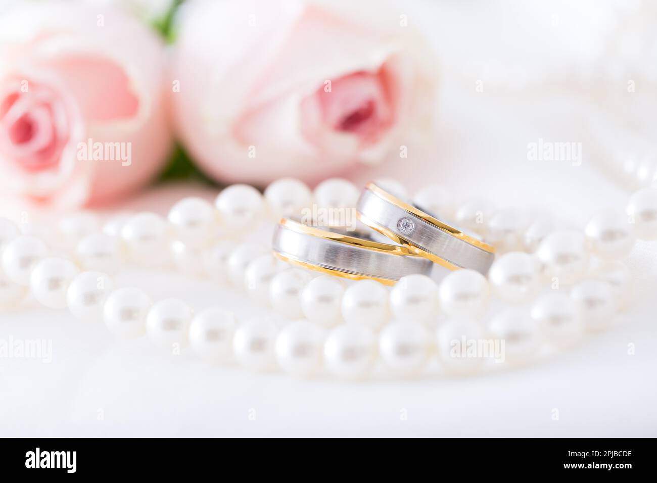 Moderne Eheringe mit Perlenkette mit rosa Rosen im Hintergrund, weicher Fokus Stockfoto