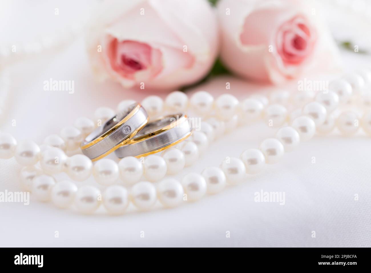 Moderne Eheringe mit Perlenkette mit rosa Rosen im Hintergrund, weicher Fokus Stockfoto