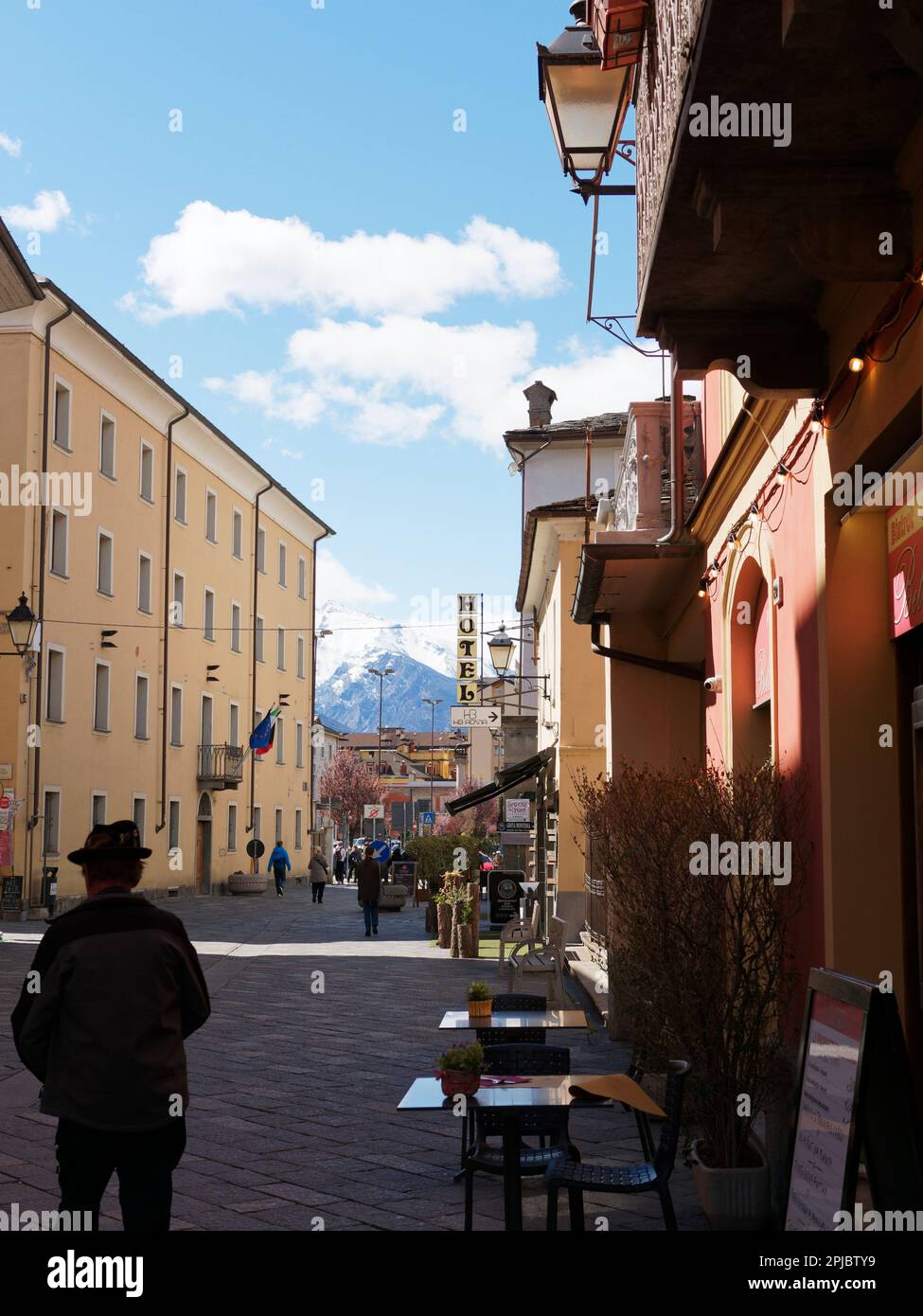 Straße in Aosta mit einem Mann mit einem Hut in Silhouette und Café/Bar Tische und Stühle im Vordergrund und einem Berg in der Ferne, Aosta Valley, Italien Stockfoto