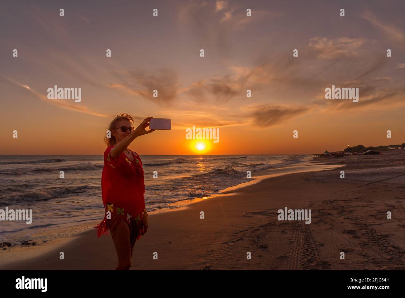 Seniorin macht ein Selfie mit einem Handy am Strand während des Sonnenuntergangs. Sommer, Urlaub, Technologiekonzept. Stockfoto