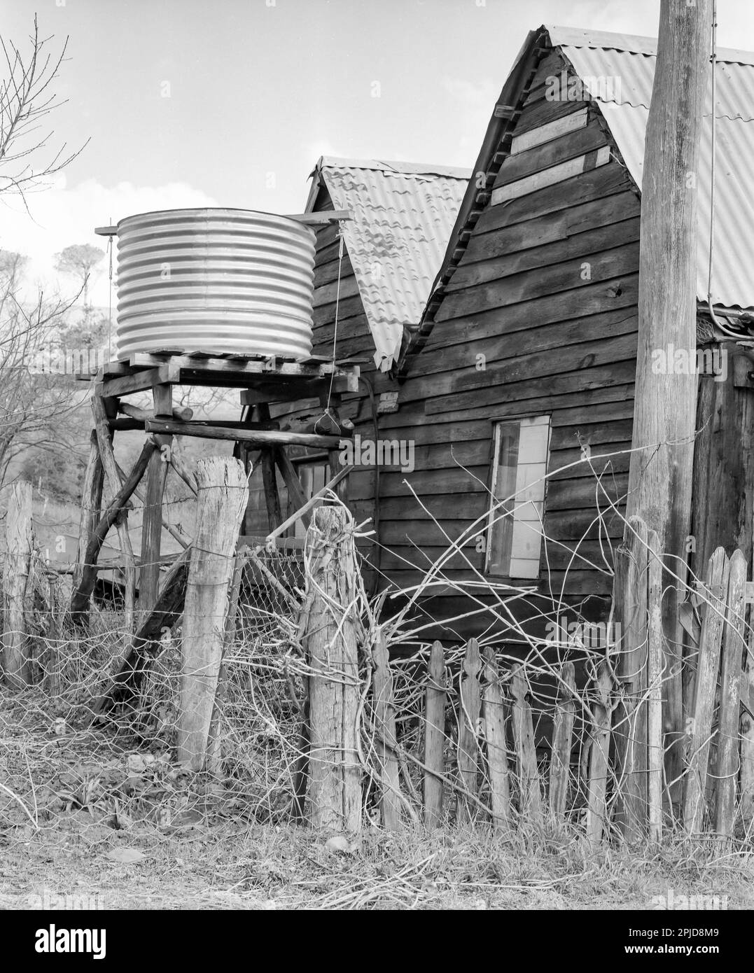 Ein 1980 schwarzweißes, mittelgroßes Foto eines Zauns aus gebrochenen alten Zaunpfählen und Hühnerdraht in der Nähe eines Hauses mit verzinktem Eisendach in der Landstadt Sofala im Zentralwesten von New South Wales, Australien. Neben dem Haus befindet sich auf einem Ständer ein verzinkter eiserner Regenwassertank, der zur Wasserversorgung des Hauses verwendet wird. Stockfoto