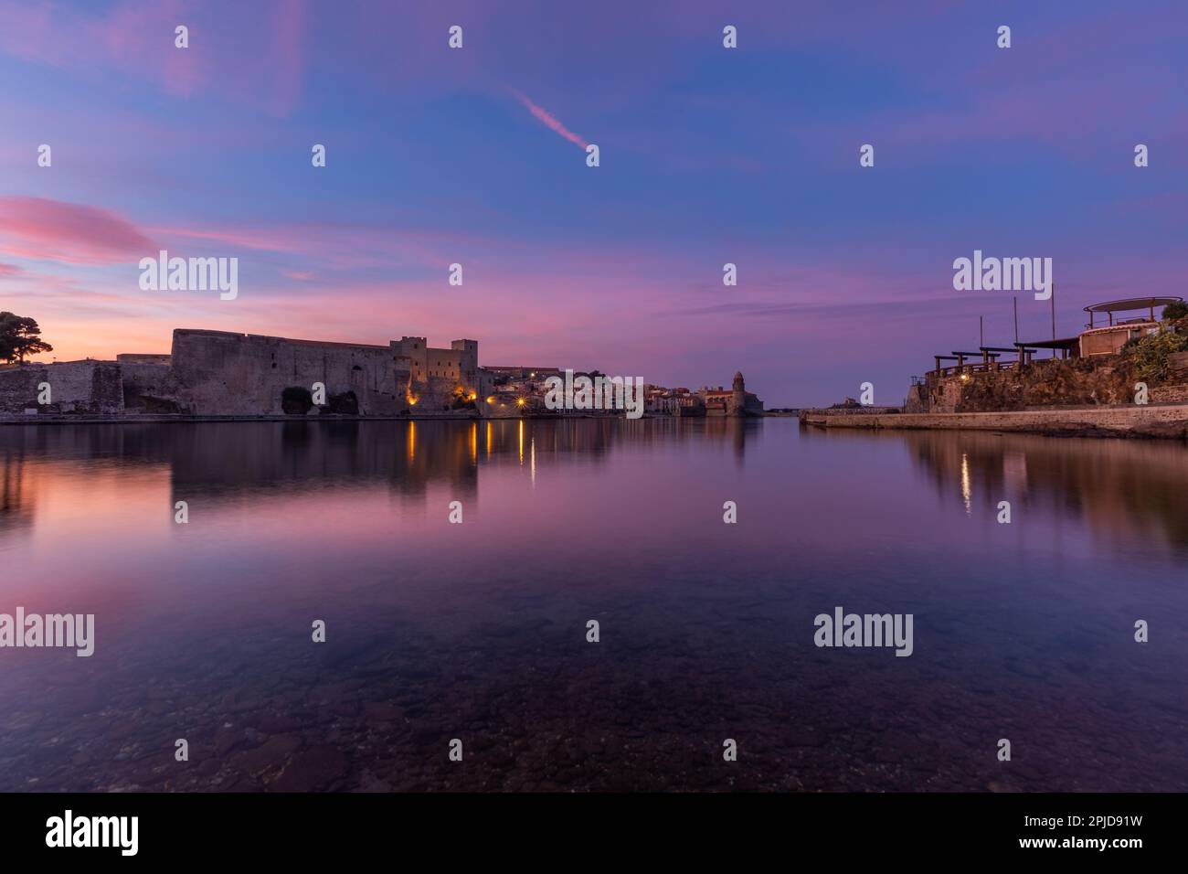 Königliche Burg von Collioure über dem stillen Wasser (Meer) in Coullioure, Frankreich nach dem Sonnenuntergang. Pink- und Violetttöne, der Himmel reflektiert sich im Wasser Stockfoto