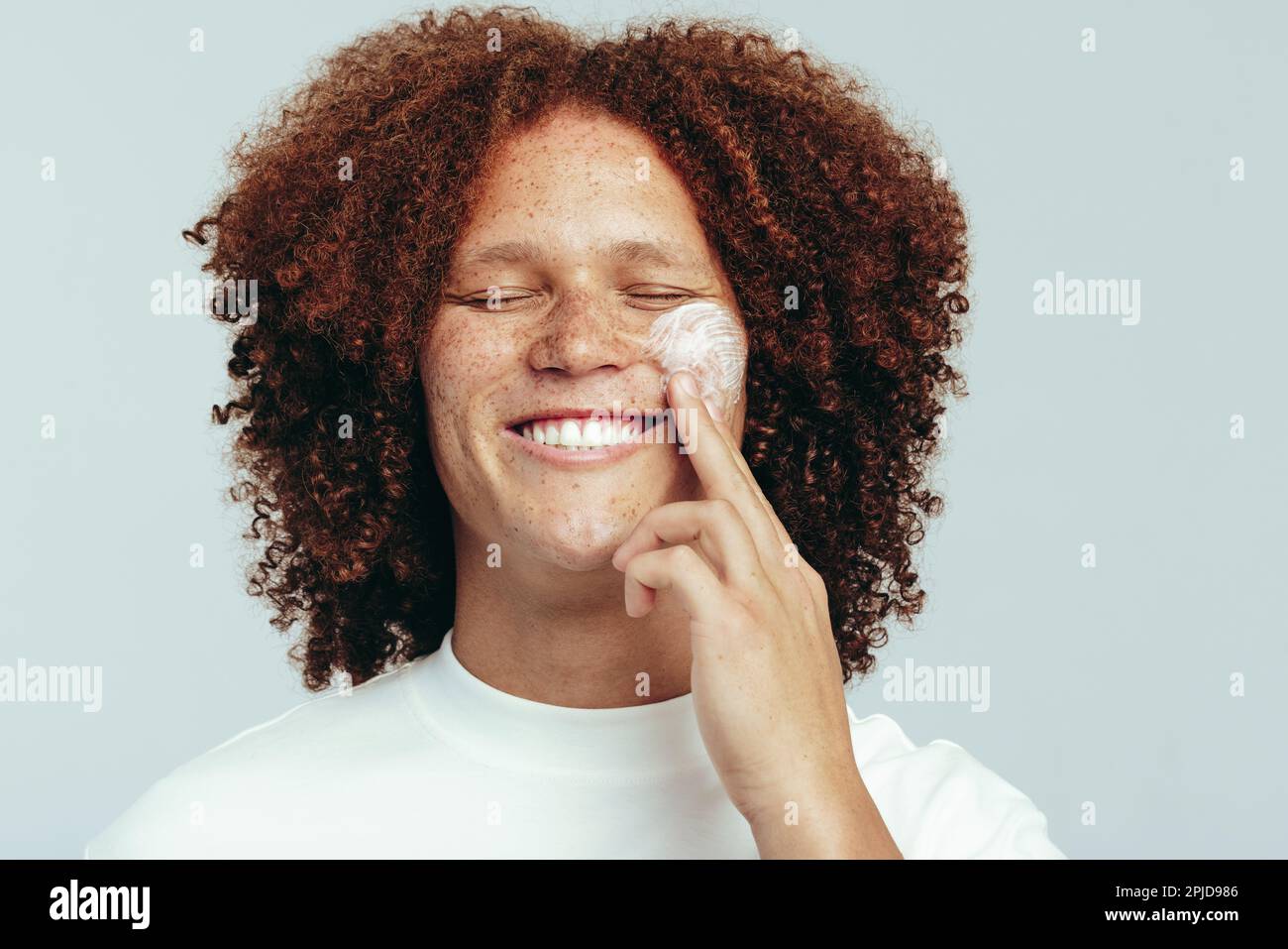 Ein Mann mit einem geschwungenen Afro, der seine Haut mit einer feuchtigkeitsspendenden Gesichtscreme pflegt. Ein hübscher junger Mann, der seine Hautpflege mit einem großen Lächeln praktiziert. Stockfoto
