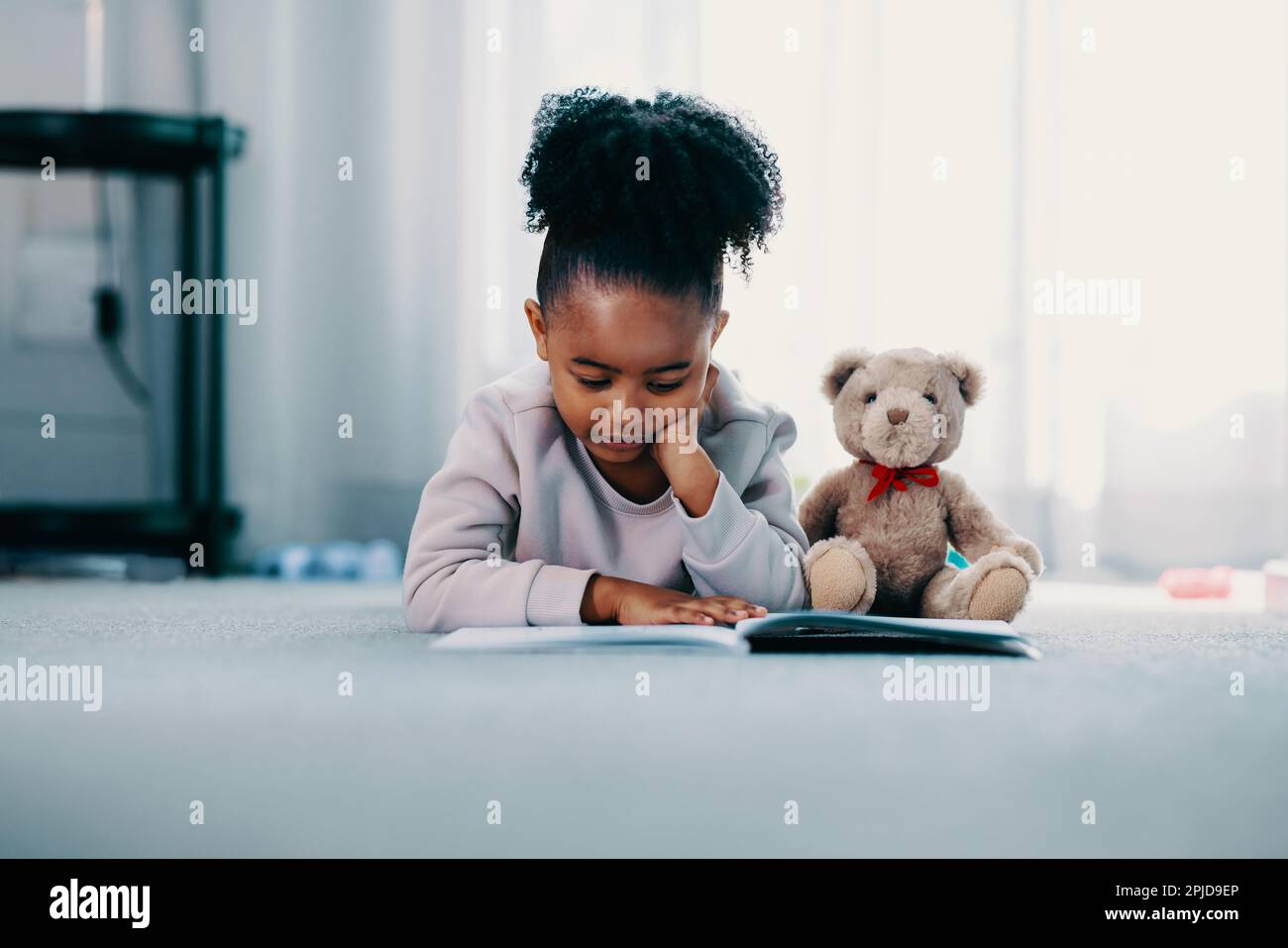 Ein Mädchen im Grundalter liegt zu Hause auf dem Boden in ihrem Schlafzimmer, sie liest ein Buch und hat ihren Teddybär neben sich. Sie konzentriert sich auf das Lernen und Stockfoto