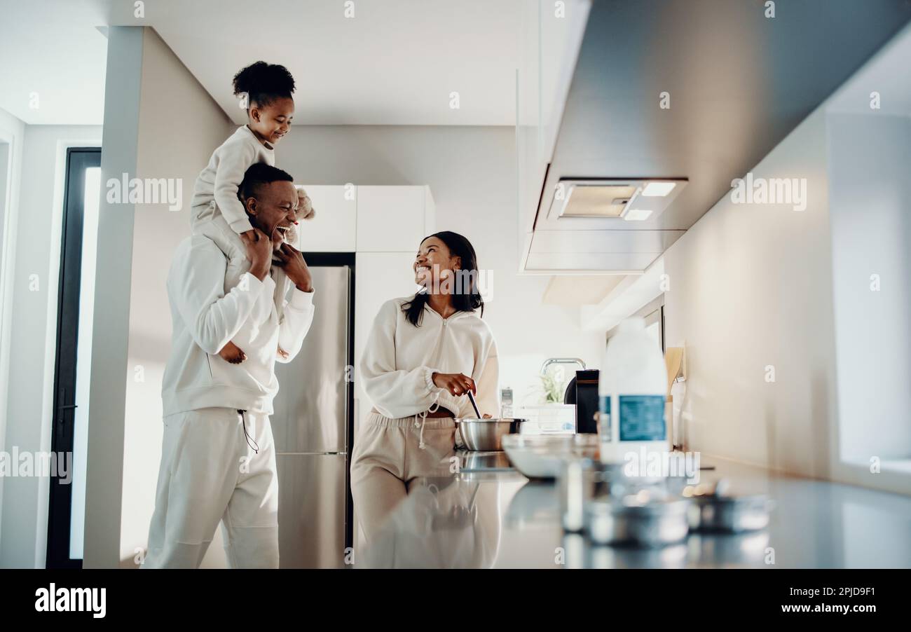 Die schwarze Familie strahlt Freude und Zusammengehörigkeit in der Küche ihres afrikanischen Heims aus. Dad trägt seine Tochter auf den Schultern, während mom kocht. Familie Stockfoto