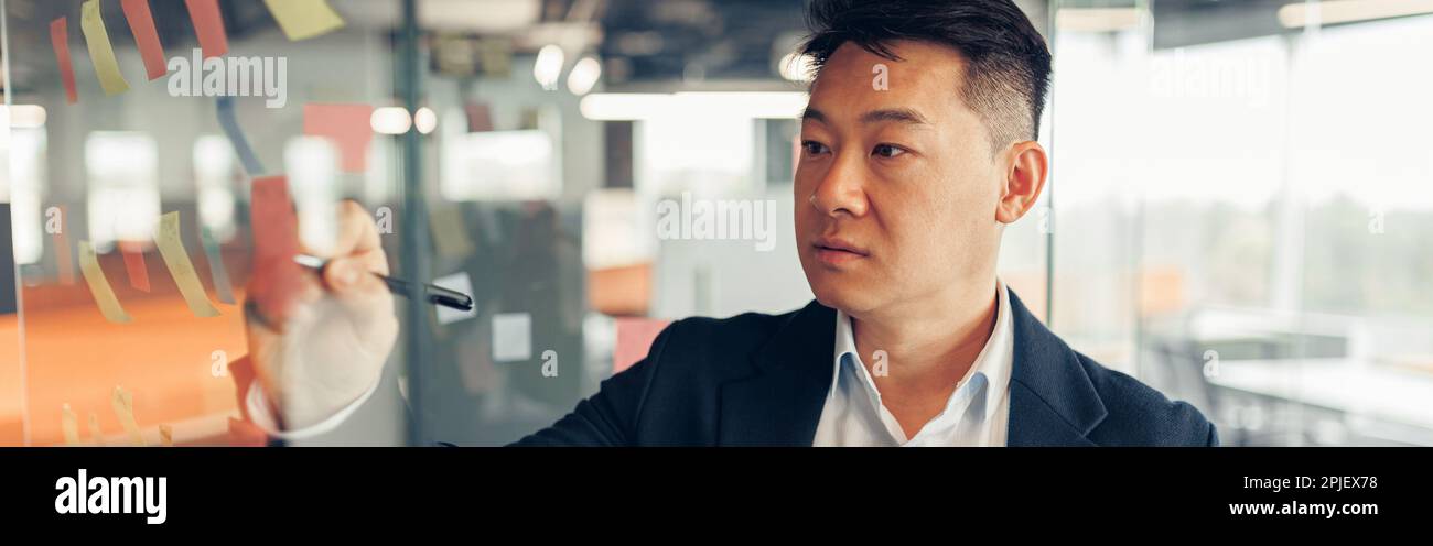 Fokussierter asiatischer Geschäftsmann, der auf Haftnotizen in der Glaswand des Büros schreibt Stockfoto