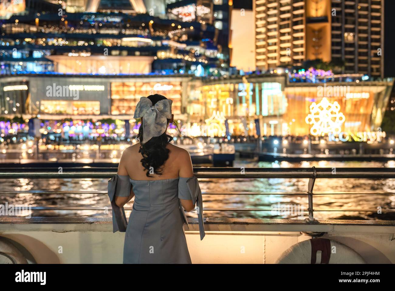 Eine schöne junge asiatische Frau im Kleid, die ein beleuchtetes Kaufhaus ansieht, das in der Nacht auf einem Festival am Flussufer glüht. Bootsfahrt mit Abendessen Stockfoto