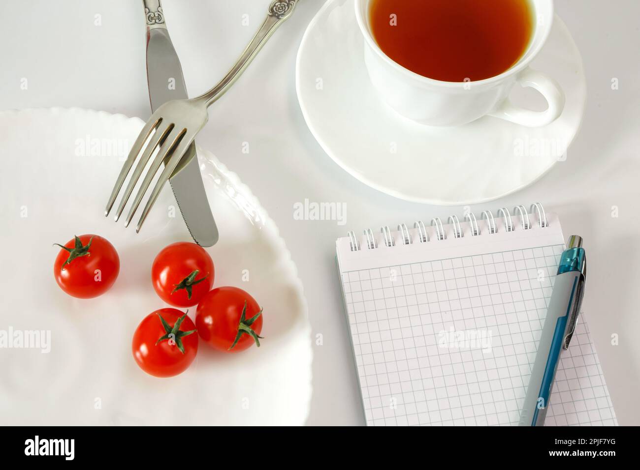 Kirschtomaten auf einem Teller mit Besteck und einer Tasse Tee. Notizbuch zum Aufzeichnen und Zählen von Kalorien für Diätkost Stockfoto