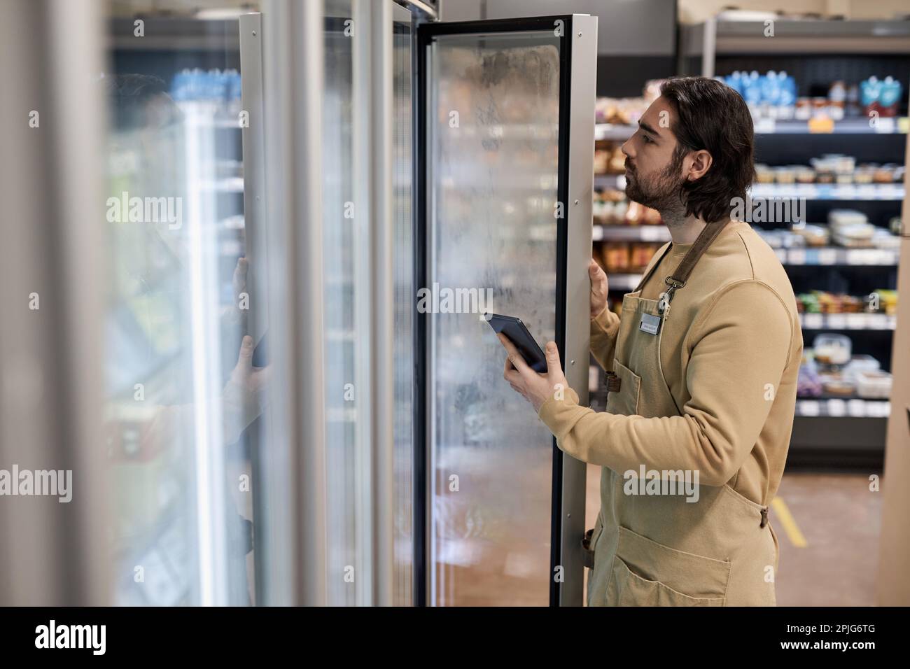 Seitliches Porträt eines männlichen Arbeiters im Supermarkt, der den Gefrierbestand inspiziert und ein digitales Tablet in der Hand hält Stockfoto