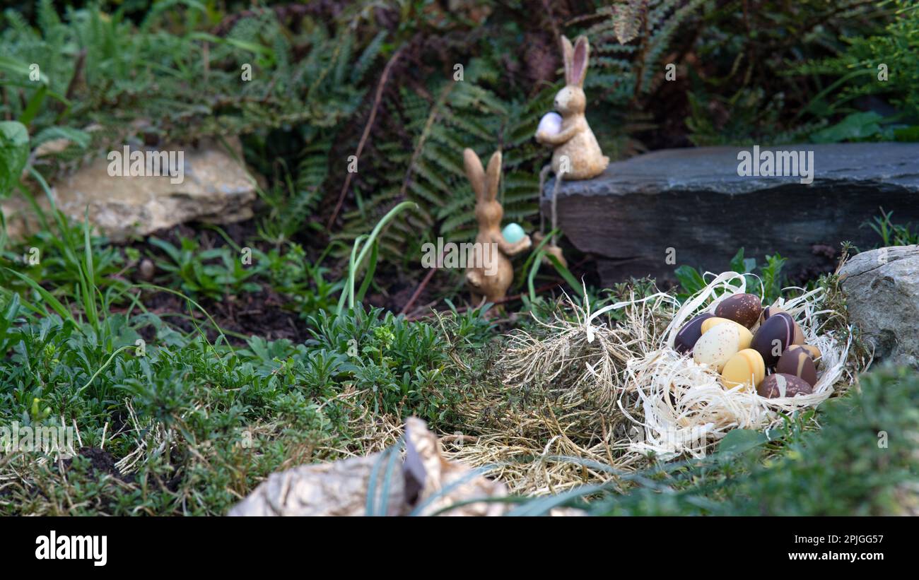Osterschokoladeneier im Nest, versteckt im Garten mit zwei kleinen Häschen im Hintergrund. Geringe Schärfentiefe Stockfoto