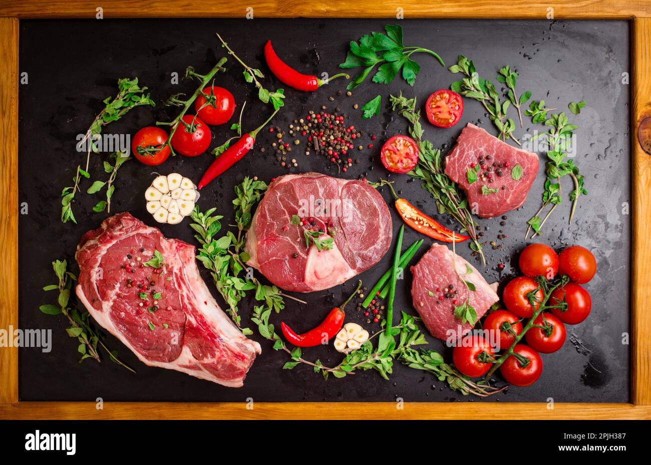 Verschiedene Steaks. Gemischtes rohes Fleisch auf schwarzem Kreidetafel mit Holzrahmen. Rib-Eye-Steak auf dem Knochen, Kalbsschenkel (Ossobuco) Stockfoto