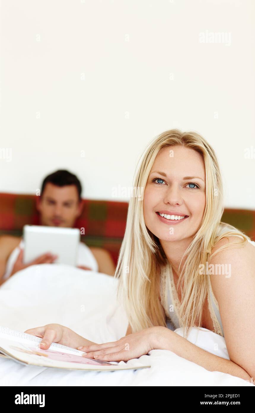 Wir machen eine Pause von unserem hektischen Lebensstil. Porträt einer jungen Frau, die im Bett liest, während ihr Mann im Hintergrund liegt. Stockfoto