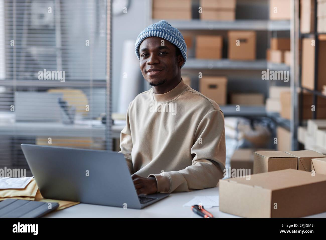 Junger afroamerikanischer Manager mit Laptop, der auf die Kamera schaut, während er am Arbeitsplatz sitzt und neue Bestellungen von Online-Kunden annimmt Stockfoto