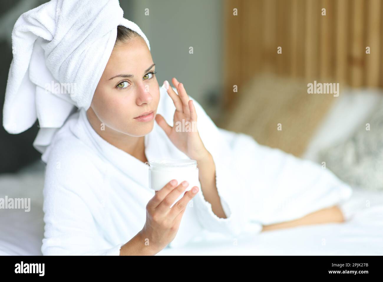 Frau, die nach dem Duschen in die Kamera schaute und ihr Gesicht mit Feuchtigkeitscreme in einem Bett zu Hause nährte Stockfoto