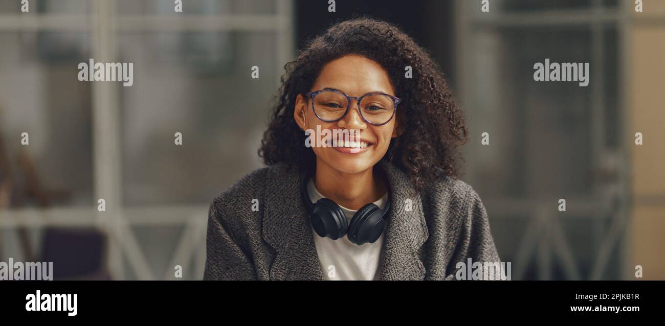 Freiberuflerin mit Kopfhörern, die vor der Kamera lächelt, während sie in moderner Zusammenarbeit mit dem Telefon sitzt Stockfoto
