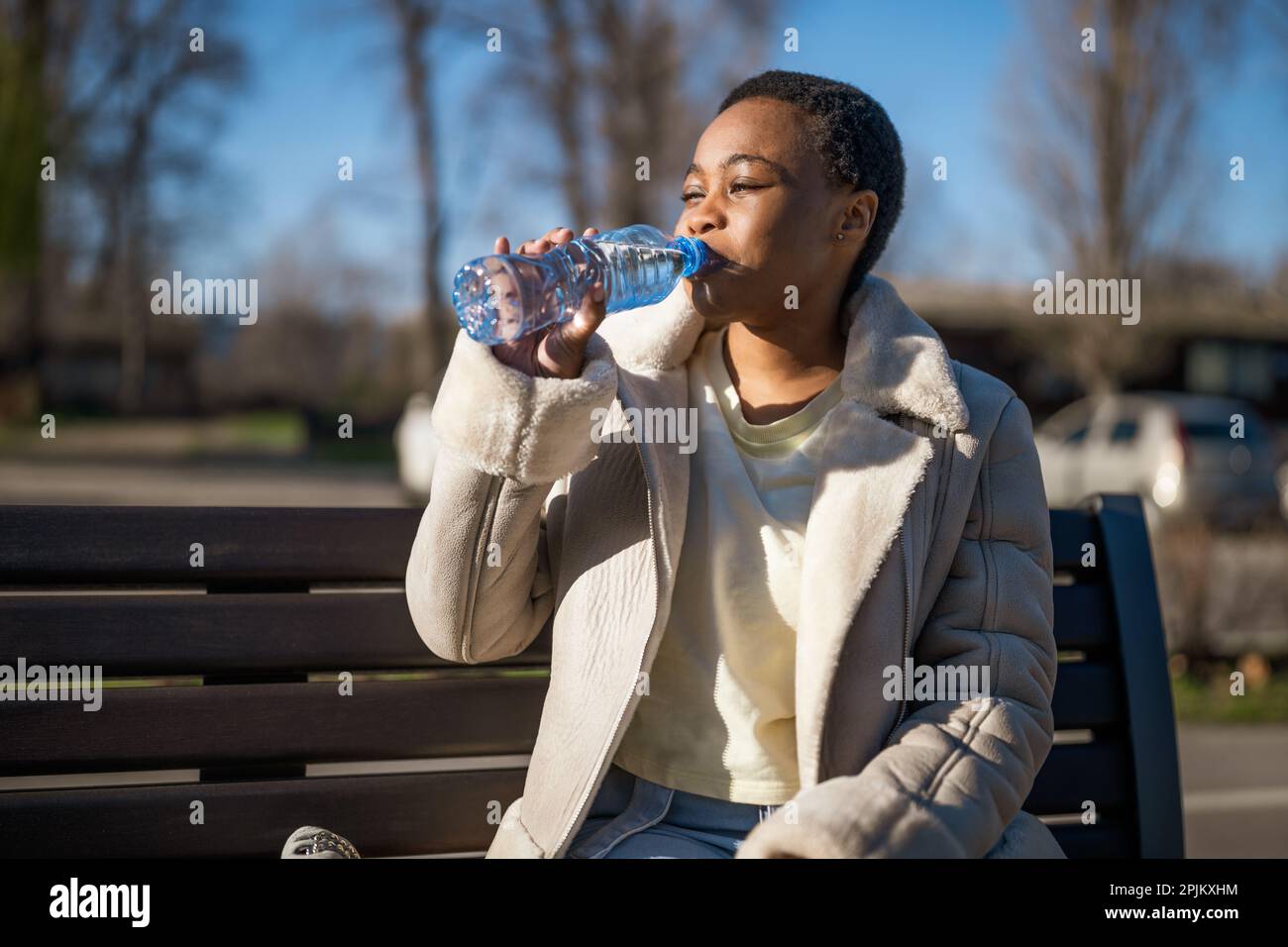Außenporträt einer glücklichen afroamerikanischen Frau am sonnigen Tag. Sie sitzt auf einer Bank auf der Straße und trinkt Wasser. Stockfoto