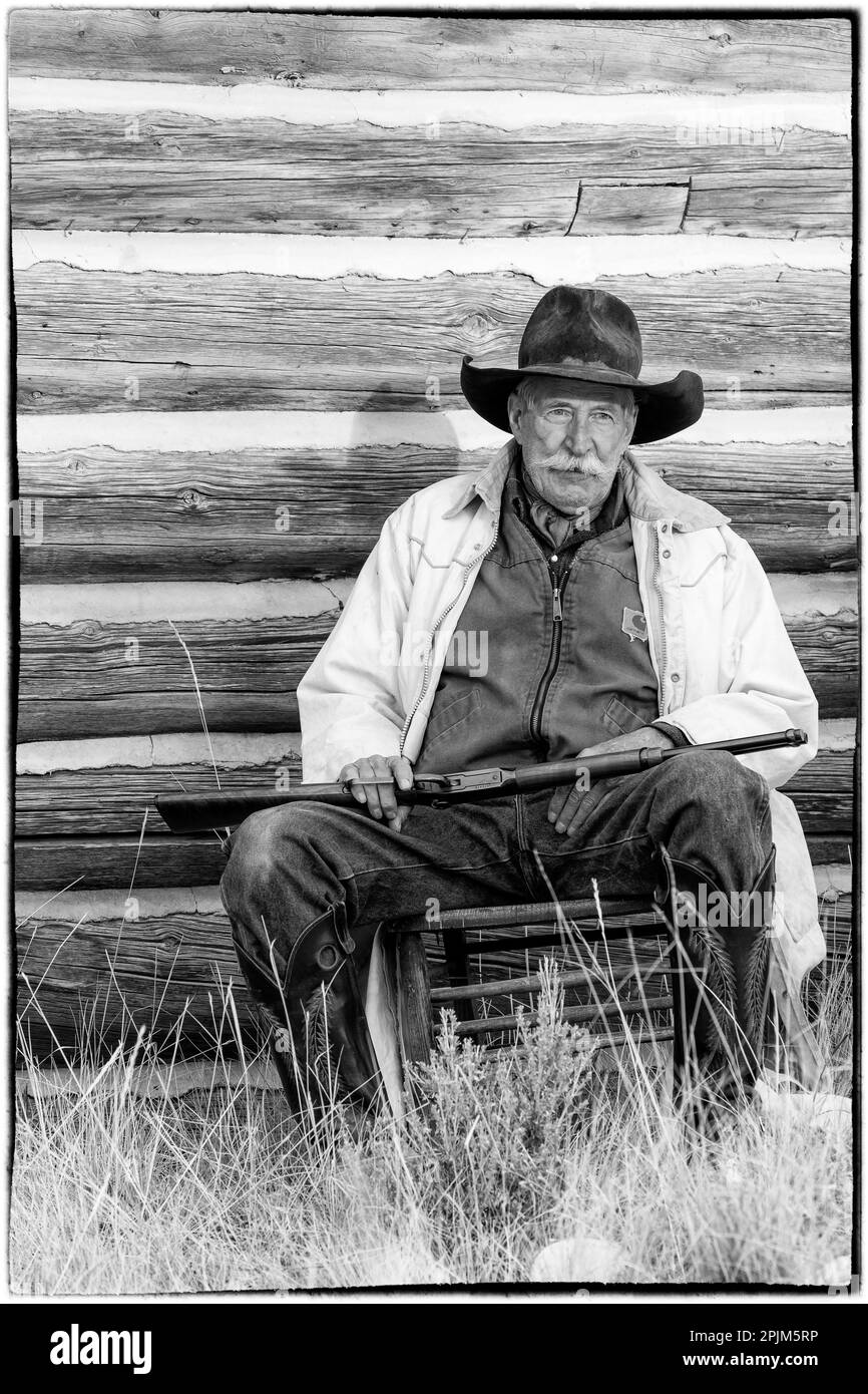 USA, Shell, Wyoming. Versteckter Cowboy der Ranch, der mit seinem Gewehr gegen die alte Blockhütte sitzt. (PR, MR) Stockfoto