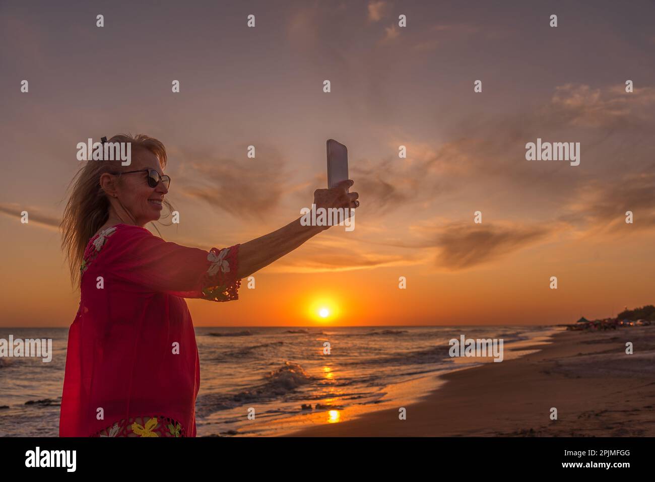 Seniorin lächelt, während sie bei Sonnenuntergang am Strand mit einem Handy Selfies macht. Sommer- und Urlaubskonzept. Stockfoto