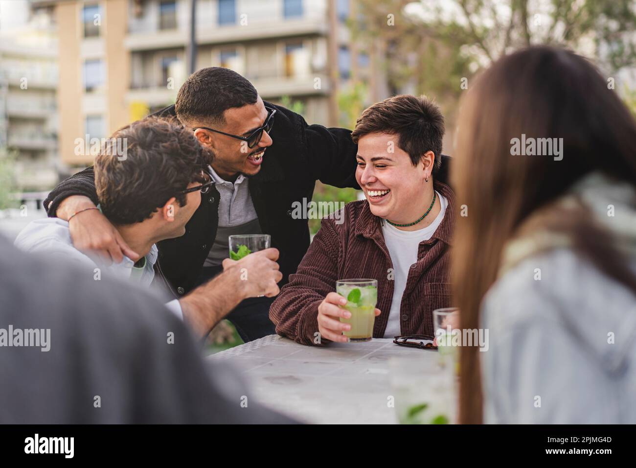 Eine vielfältige Gruppe von Freunden, darunter eine nicht binäre Person in Übergröße und ein weißer Mann, genießen Mojitos in einem Pub im Freien, während sie gerne einen gemischten begrüßen Stockfoto