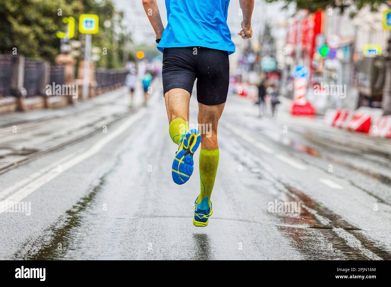 Rücken Sie männlichen Sportler in Kompressionssocken zurück, laufen Sie Marathon in der Stadt, Sommerrennen, joggen auf nassem Asphalt nach Regen Stockfoto