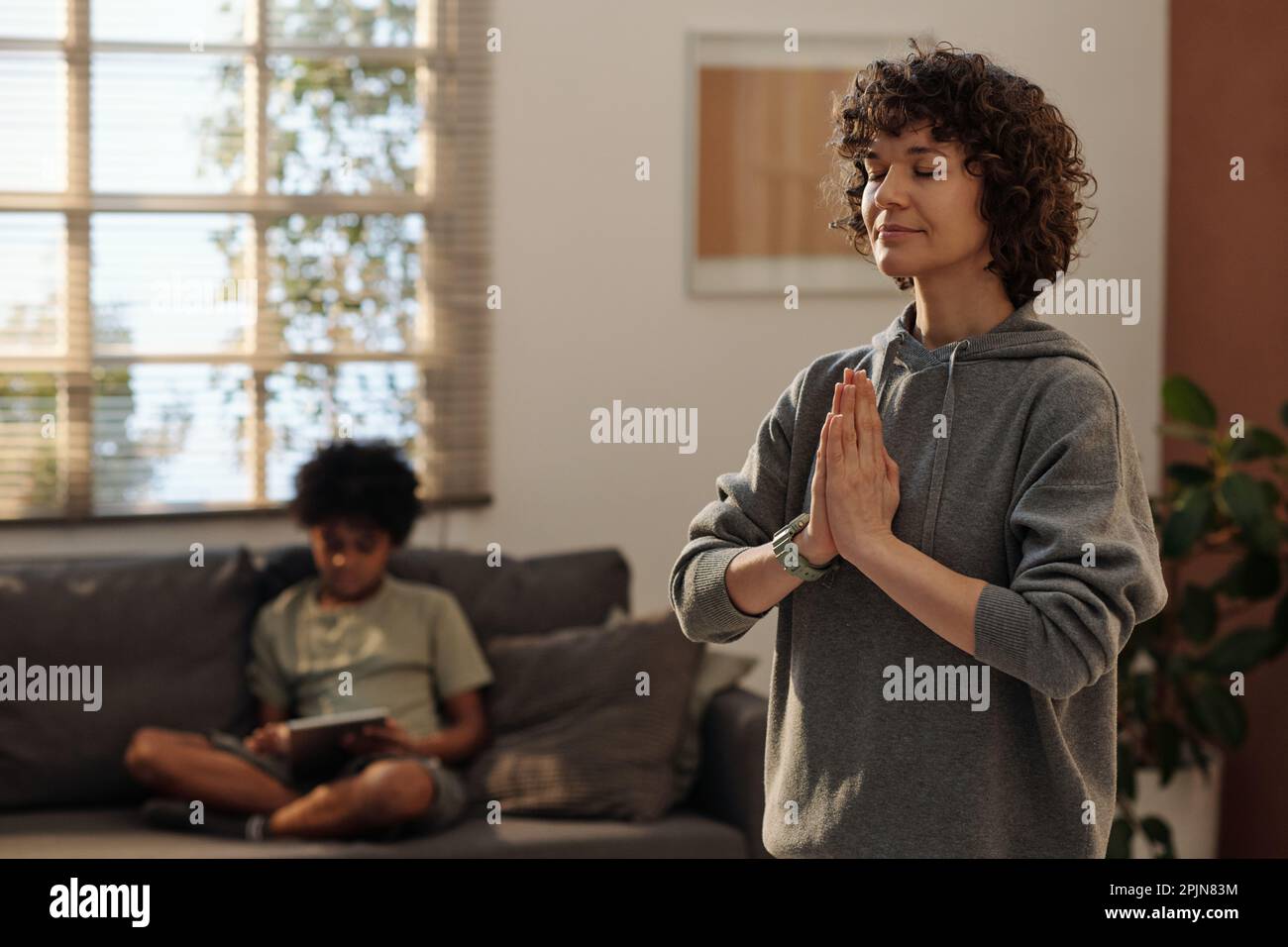 Junge, entspannte Frau in grauem Hoodie, die während des Yoga-Trainings die Hände zusammenhält, während sie zu Hause vor der Kamera steht Stockfoto