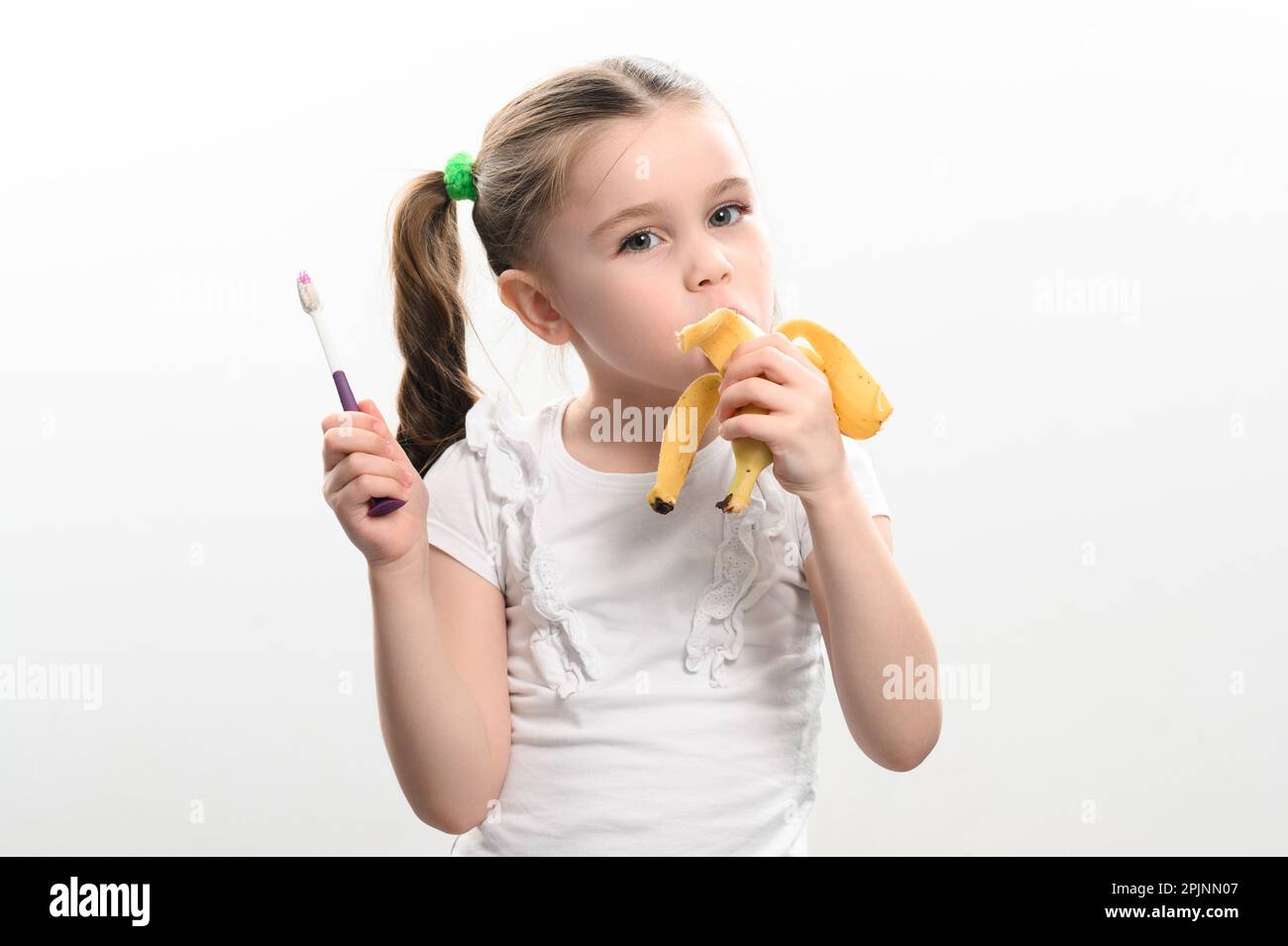 Ein kleines Mädchen isst eine Banane und hält eine Zahnbürste in der Hand, Studiofoto auf weißem Hintergrund, eine Banane hilft gegen Karies, eine Idee für Zahnärzte Stockfoto