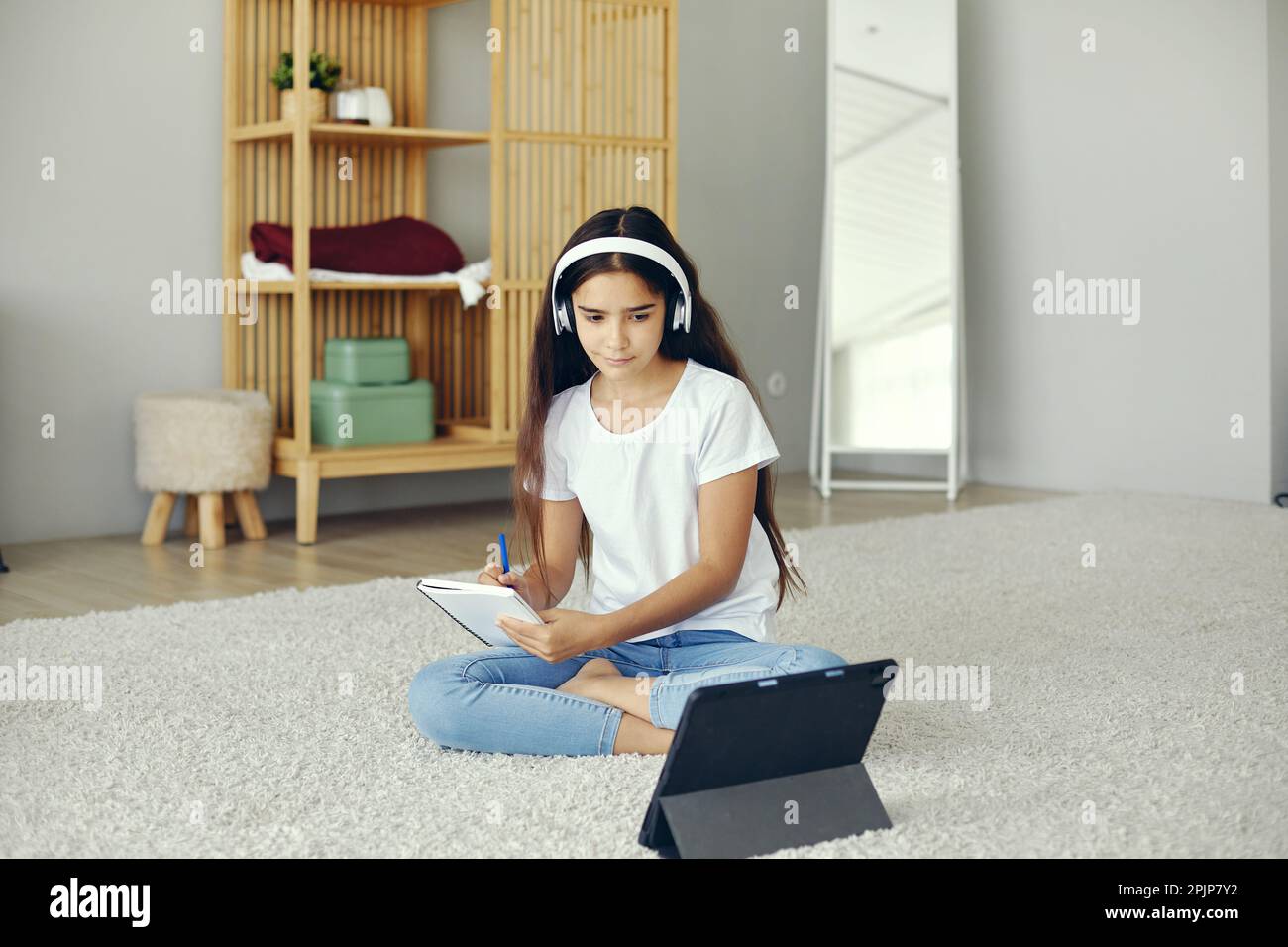 Hübsches Mädchen im Alter von 12s Jahren trägt kabellose Kopfhörer, sitzt auf dem Boden, hört Audiokurse, lernt zu Hause und lernt mit einem digitalen Tablet. Moderne Technik f Stockfoto
