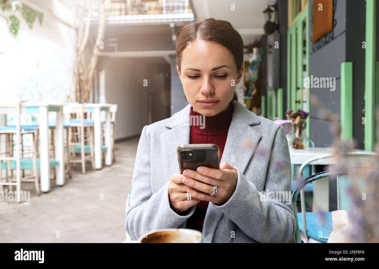 Eine Frau, die mit ihrem Handy SMS schreibt, sitzt in einem Café im Freien. Stockfoto