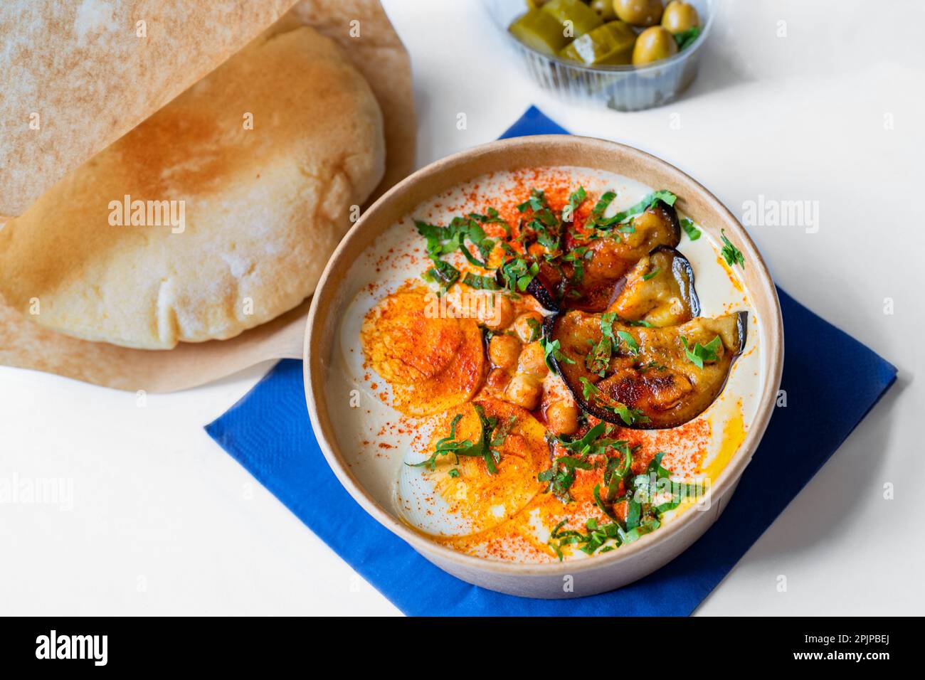 Schüssel mit Hummus, in Scheiben gekochtem Ei, gerösteten Auberginen und grünem Shoot, Brot und Oliven auf blauer Serviette auf weißem Hintergrund. Stockfoto