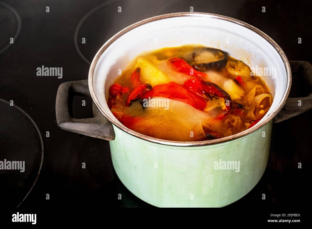 Kochende und sprudelnde Suppe aus rotem und gelbem Pfeffer und Tomate in grünem Topf auf schwarzem Keramikkochfeld. Zubereitung der Suppe aus Pfeffer und Tomate. Stockfoto