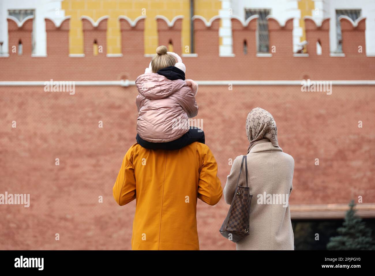 Familienpaar mit einem Kind, das auf dem Roten Platz in Moskau auf dem Hintergrund der Kremlmauer steht. Das kleine Mädchen sitzt auf den Schultern des Vaters, Eltern in Spring City Stockfoto