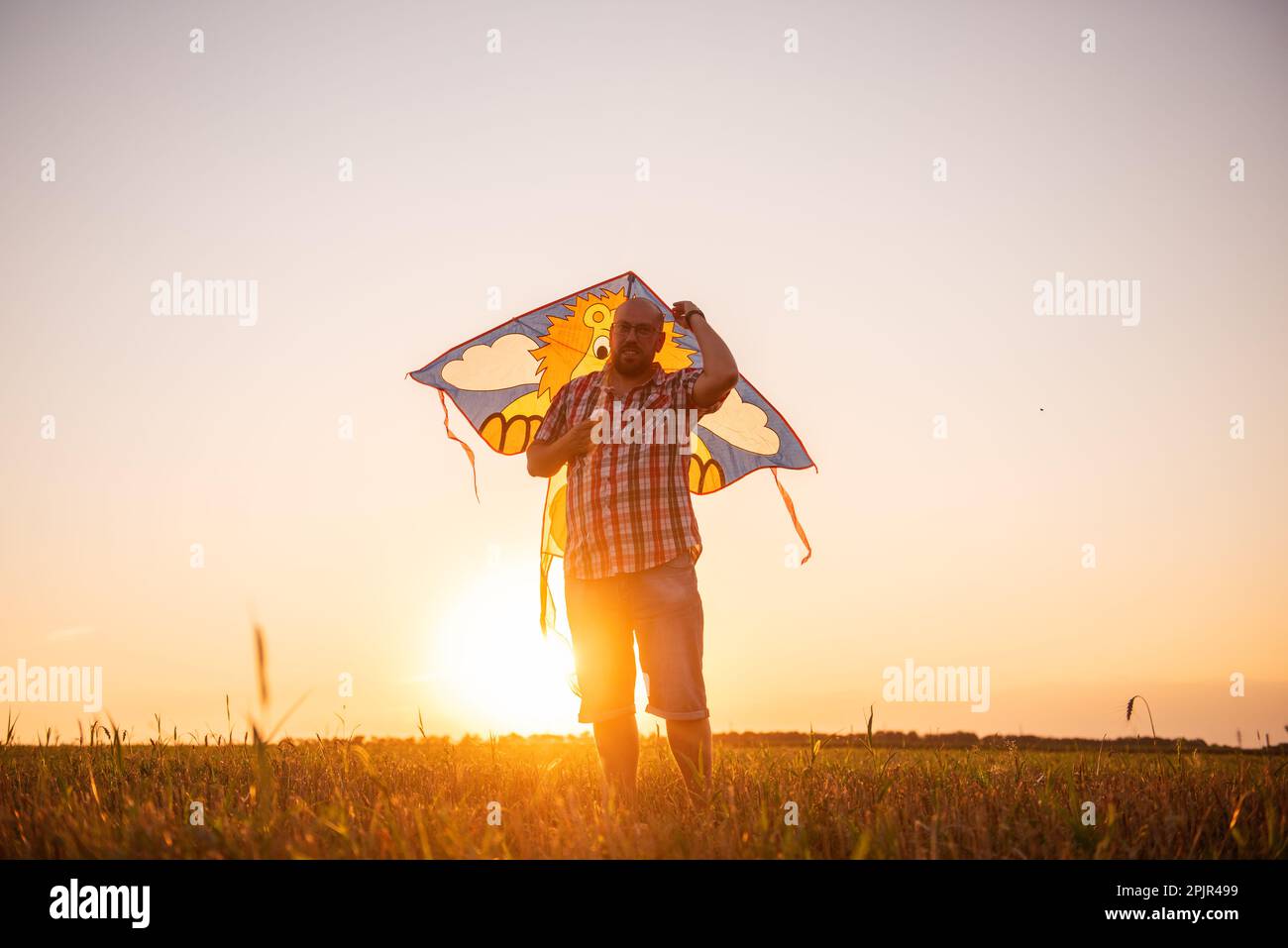 In den Sonnenstrahlen des Sonnenuntergangs, kahlköpfiger Mann mit Brille und Drachen auf dem Feld. Vater spielt mit Kindern in ländlichen Gegenden. Blick durch die Ohren des m. Stockfoto