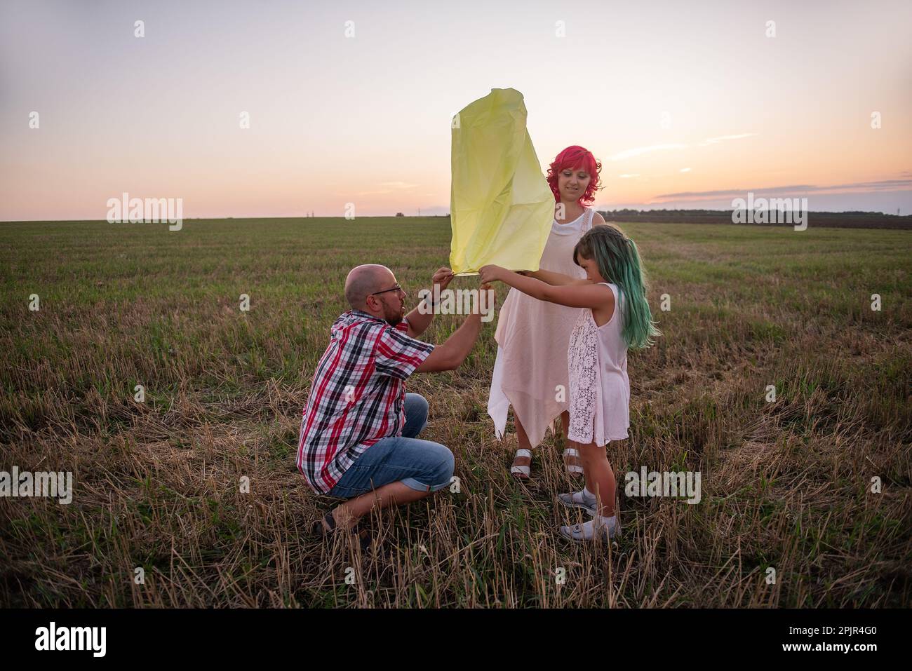 Diversity-Familie, Mutter Vater Tochter zündet eine gelbe Papierlaterne am Himmel bei Sonnenuntergang auf dem Feld an. Teamarbeit. Wünsche machen. Das Konzept der Hoffnung, Fai Stockfoto