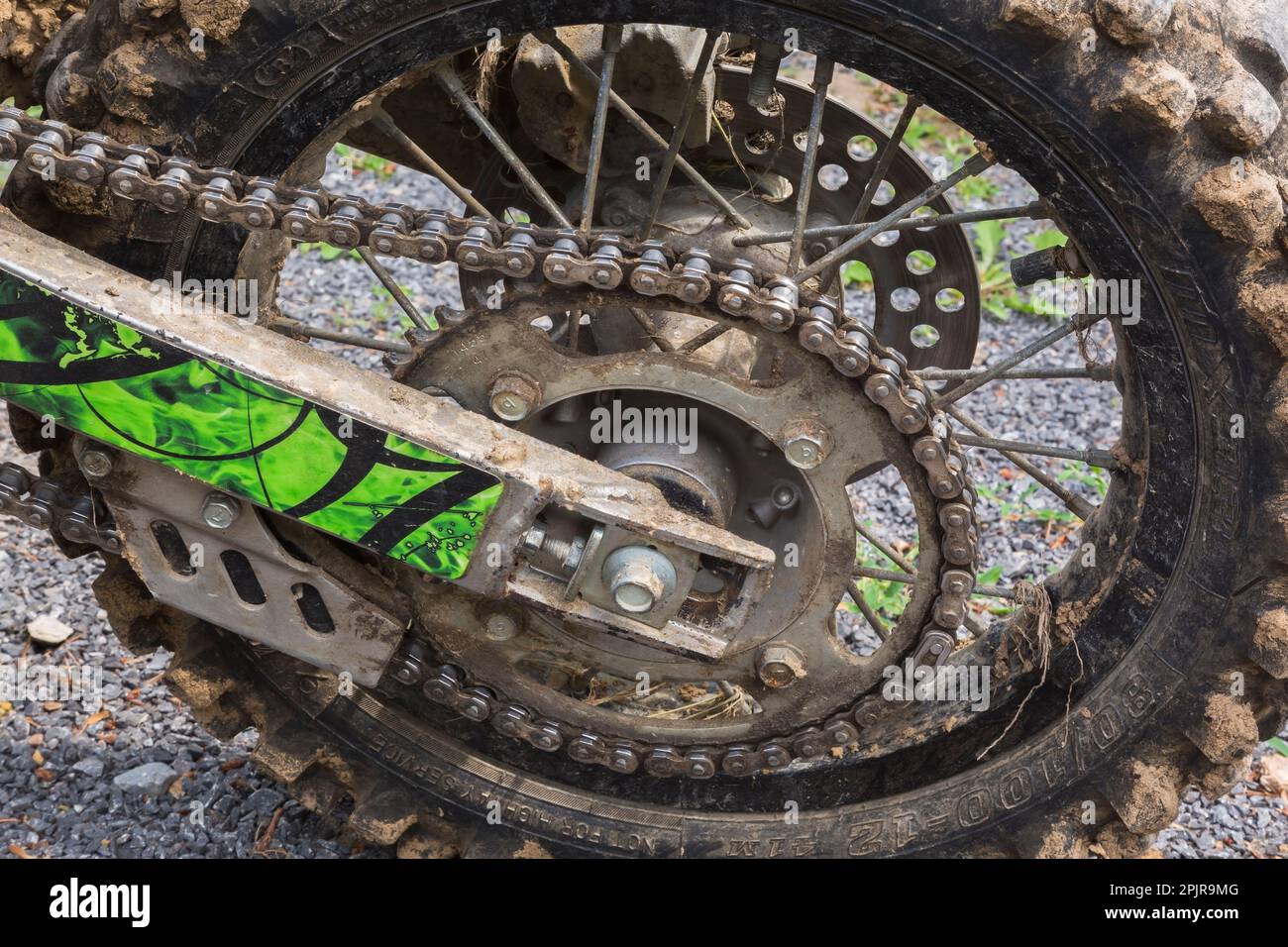 Kette und schwarzer hinterer Gummireifen, der mit getrocknetem Schlamm auf einem auf Schotterstraßen geparkten Motorrad bedeckt ist. Stockfoto