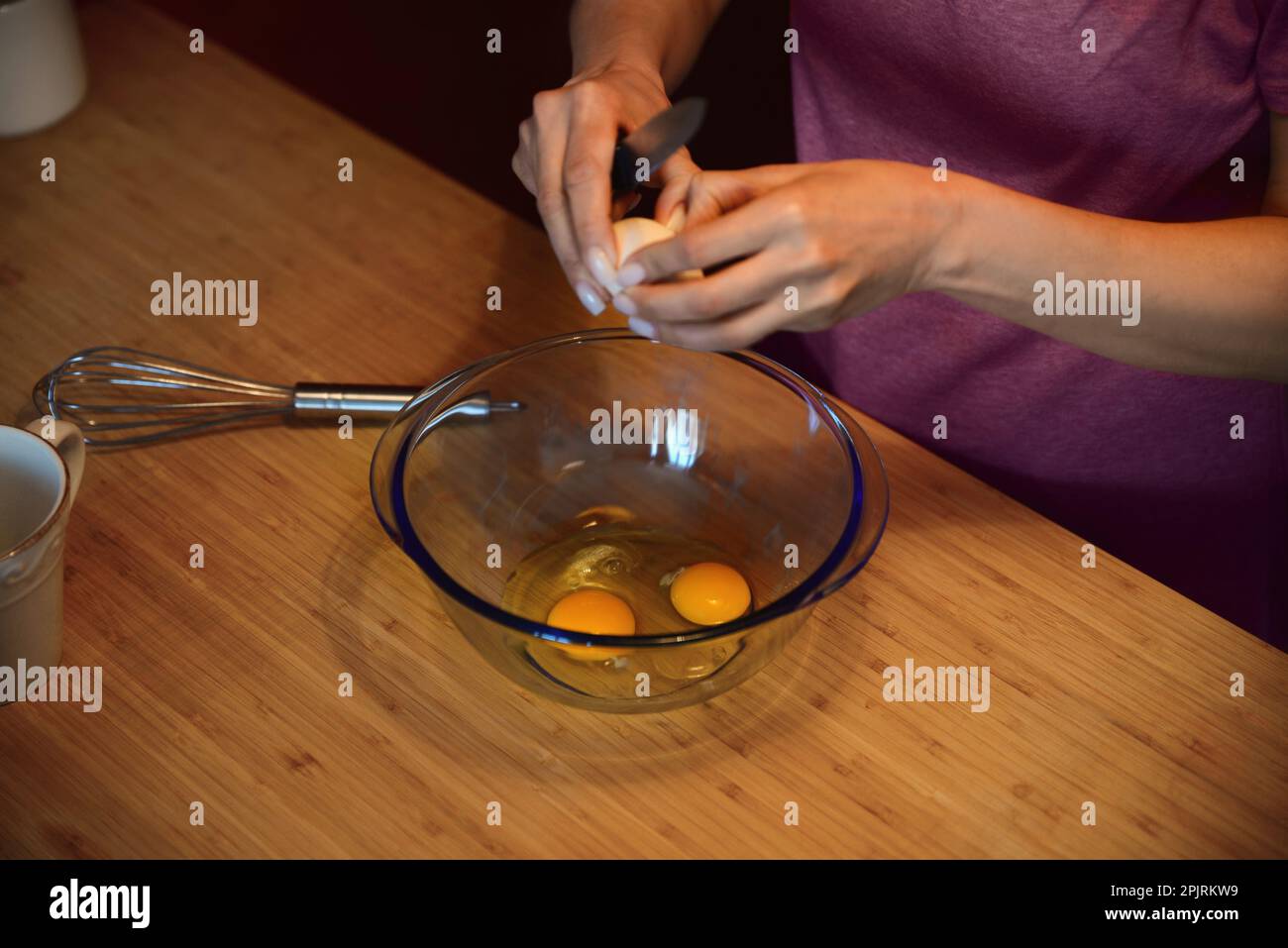 Eine Frau zerbricht Eier für Crepe-Teig in einer Glasschüssel auf einem Holztisch, Nahaufnahme Stockfoto