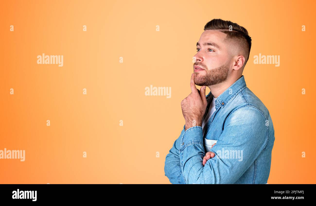 Nachdenklicher junger, gutaussehender Mann mit Hand am Kinn, Profilporträt, das Pläne macht, orangefarbener, leerer Kopierraum-Hintergrund. Gedanken- und Ideenbegriff Stockfoto