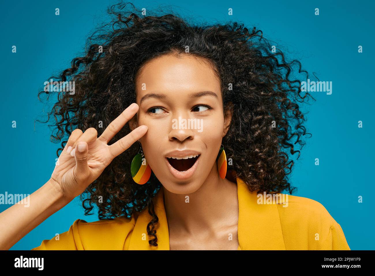 Überraschende Emotionen auf dem Gesicht einer afroamerikanischen Frau mit zwei Fingern, die ein Siegeszeichen macht, sie kann ihr Glück nicht glauben, auf blauem Hintergrund Stockfoto