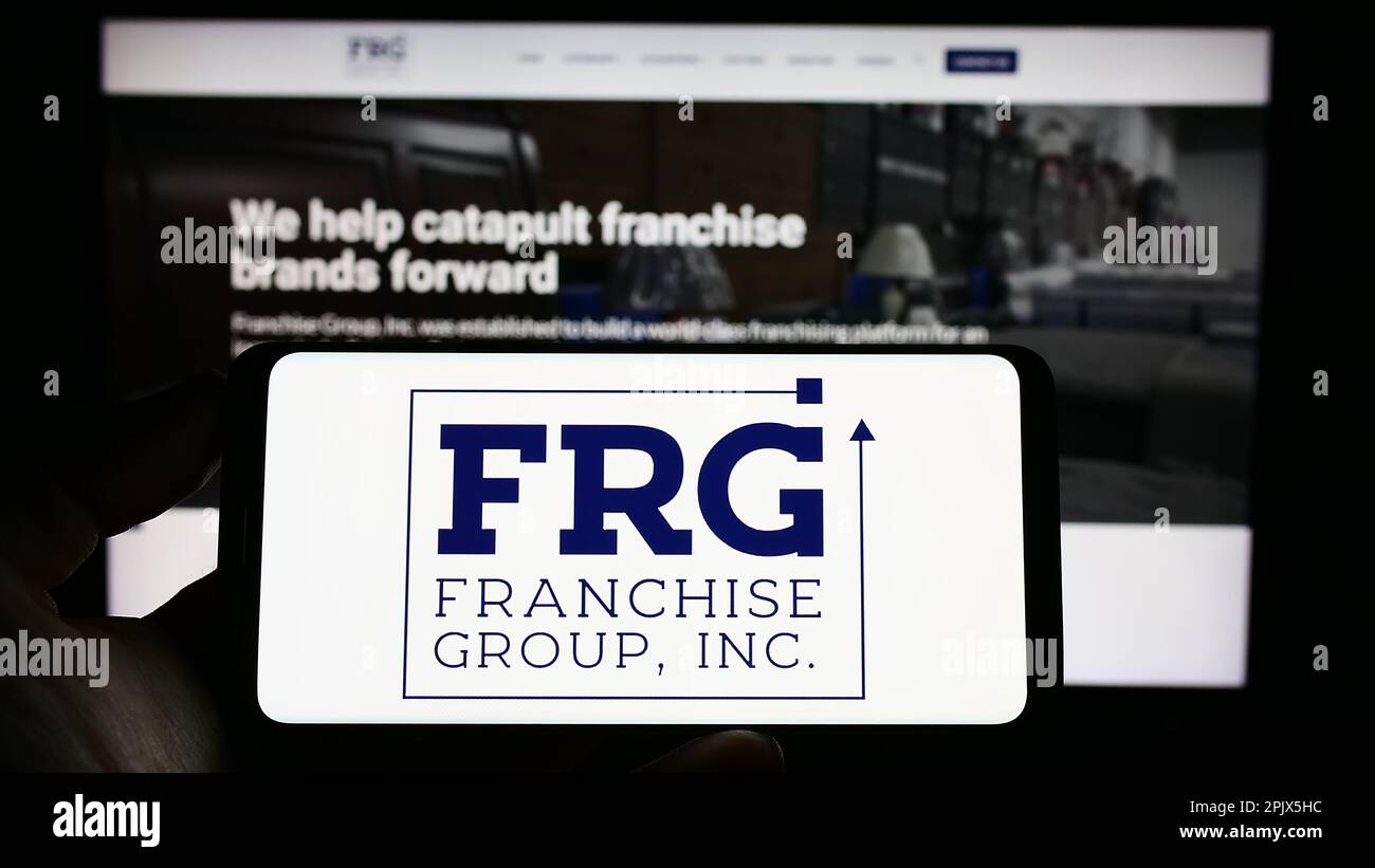 Person, die ein Mobiltelefon mit dem Logo der US-amerikanischen Franchise Group Inc. Besitzt (FRG) auf dem Bildschirm vor der Webseite des Unternehmens. Konzentrieren Sie sich auf das Display des Telefons. Stockfoto