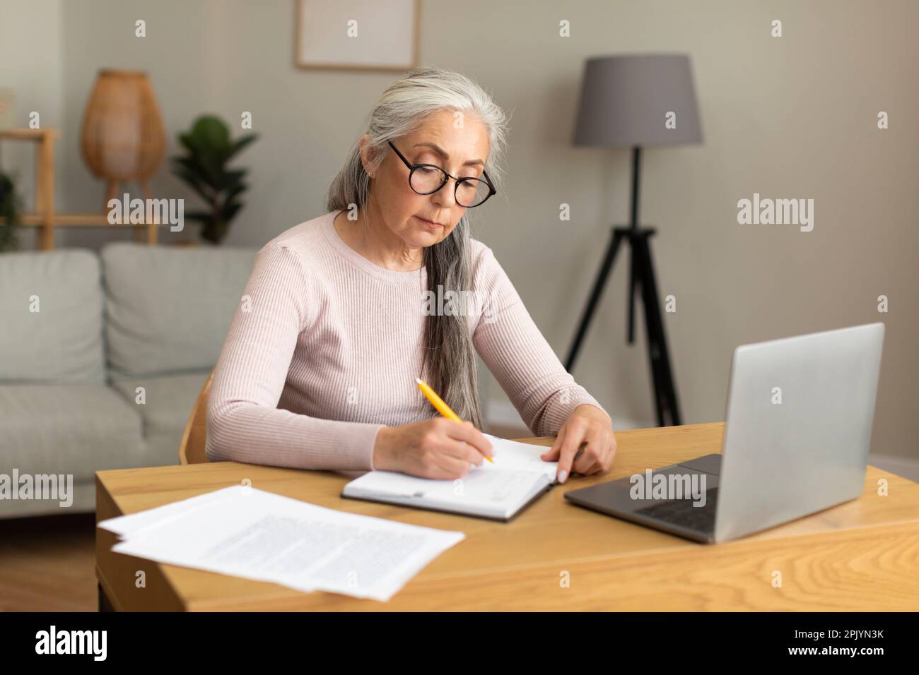 Konzentrierte, fleißige, weiße, alte Dame mit Brille macht Notizen am Tisch mit Computer Stockfoto