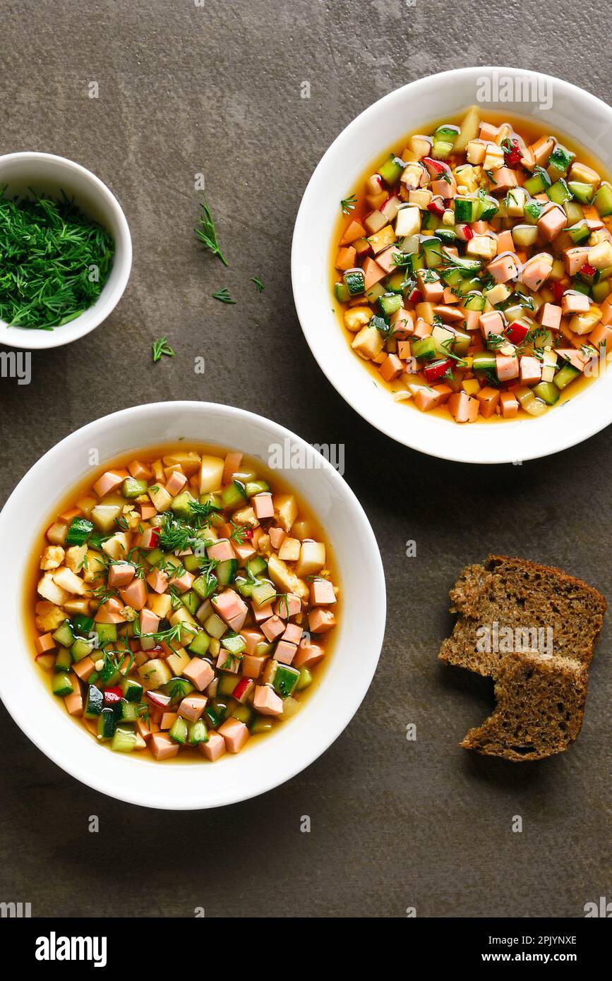 Kalte Okroshka-Suppe mit Wurst, Gemüse und Kvass in Schüsseln auf braunem Hintergrund. Draufsicht, flach liegend Stockfoto