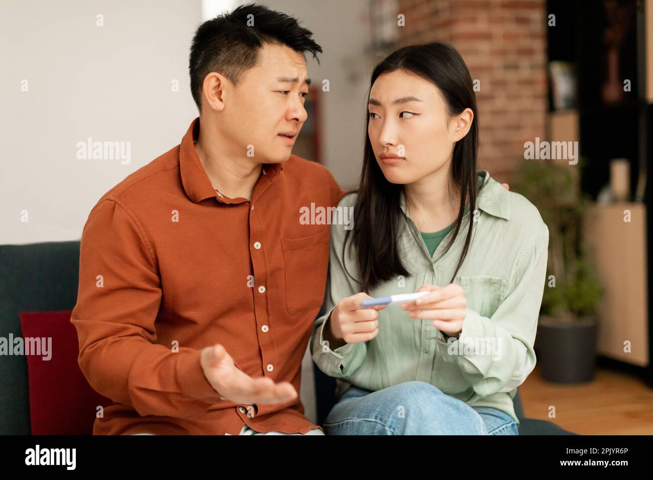 Deprimiert traurig asiatischer Ehemann und Ehefrau aufgebracht wegen negativem Schwangerschaftstest Ergebnis, sitzend auf Sofa im Wohnzimmer Innenraum Stockfoto