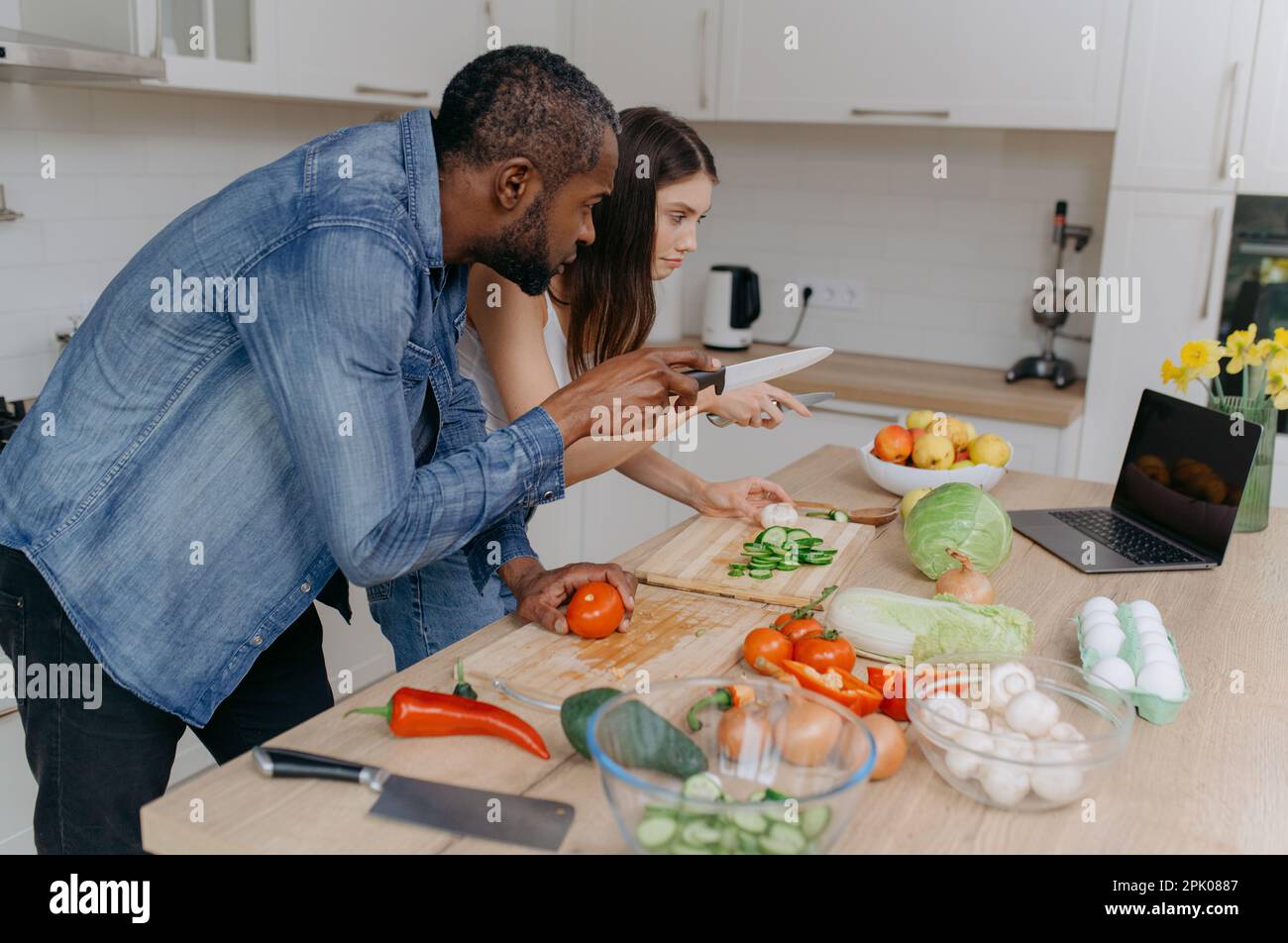 Ein verheiratetes Paar kocht zusammen mit einem Laptop und sucht im Internet nach Rezepten. Ein Afroamerikaner und seine Frau bereiten einen Salat im Set zu Stockfoto