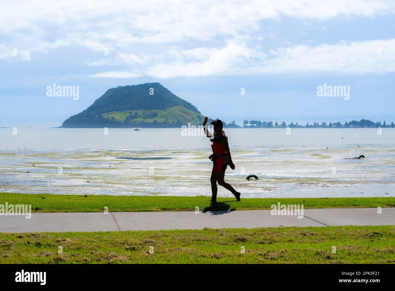 Tauranga Neuseeland - 2 2023. April; Frau in Silhouette spaziert zügig am Ufer entlang mit dem Wahrzeichen Mount Maunganui im Hintergrund. Stockfoto