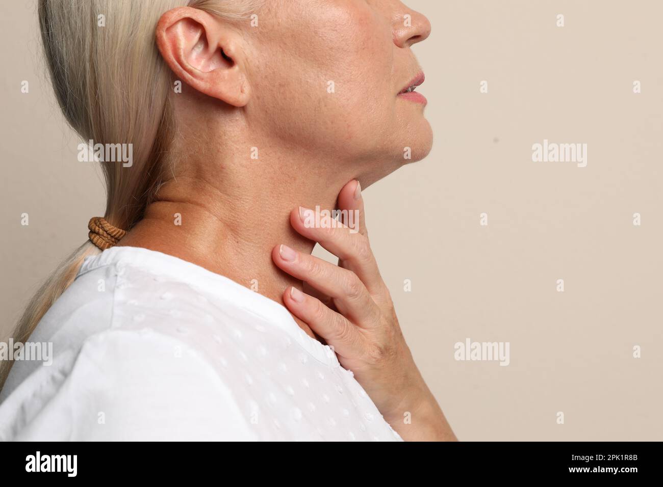 Reife Frau, die Schilddrüsen-Selbstuntersuchung bei beigefarbener Hintergrundnaht macht Stockfoto