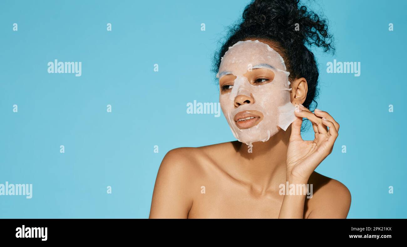 Gesichtshydratisierungs- und Verjüngungsverfahren. Afroamerikanische Frauenporträt mit feuchtigkeitsspendender Hautpflegemaske auf blauem Hintergrund, hohe Qualität Stockfoto