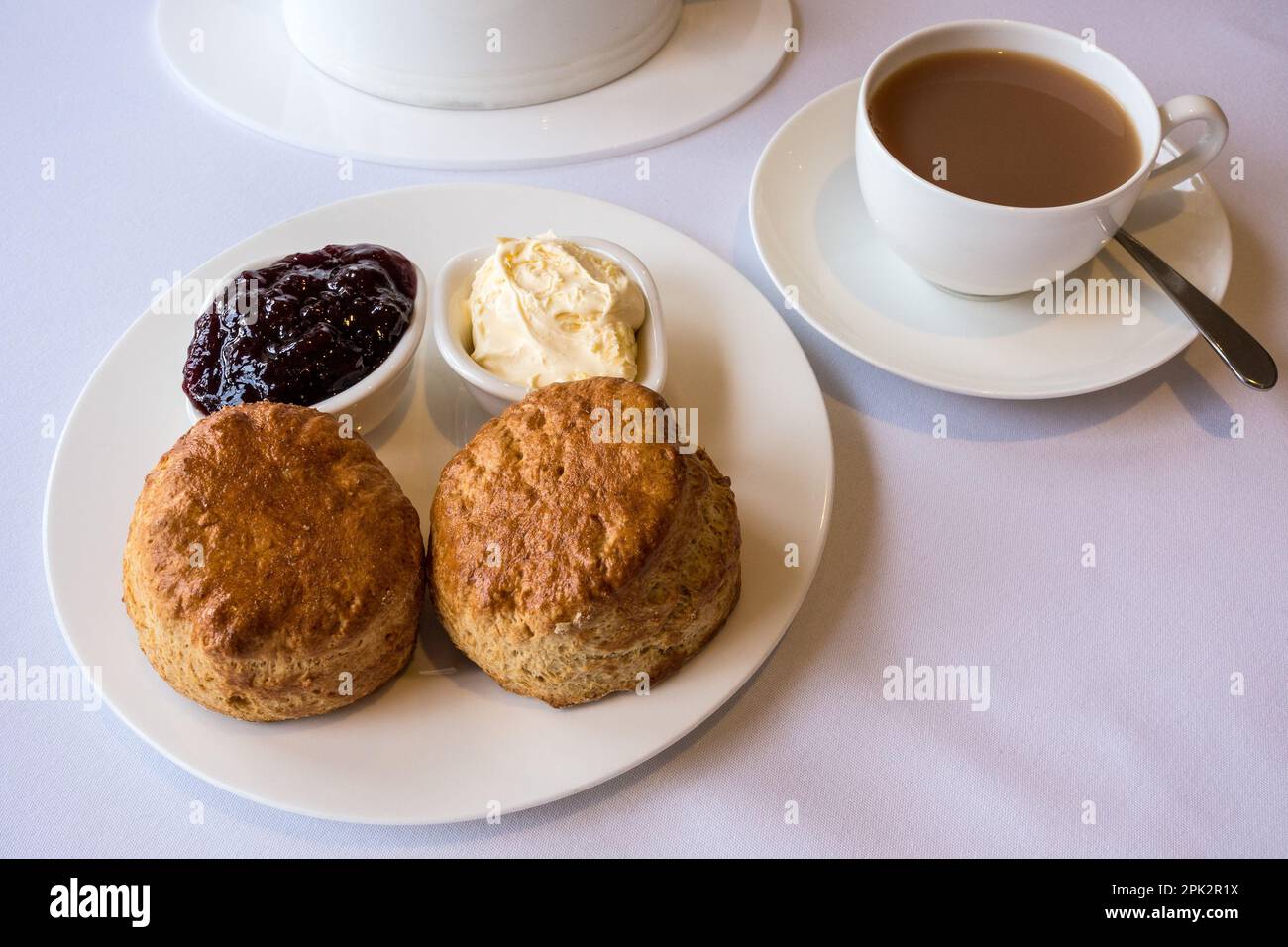 Cream Tea mit zwei frisch gebackenen einfachen Scones mit Geschirr aus Clotted Cream und Marmelade sowie einer Tasse Tee, serviert auf sauberem weißem Geschirr und Tischdecke. Stockfoto