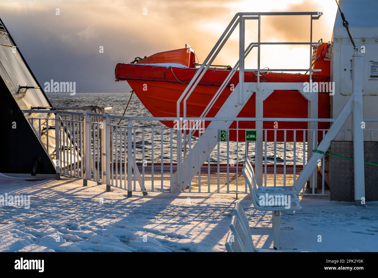 Teil eines orangefarbenen Rettungsboots auf Schnee und eisbedeckten Fähren-Deck, dramatischer und sonniger Himmel bei Sonnenuntergang. Von Moskenes nach Bodø, Lofoten, Norwegen. Stockfoto